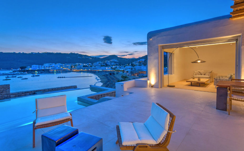 Terrace lounge chairs and pool at dusk with the lights of Platis Gialos bay beyond, Villa Laina, Mykonos