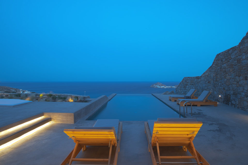 Infinity pool and wooden sun loungers at dusk with stone wall and sea view at Villa Pierre, Kalafatis, Mykonos