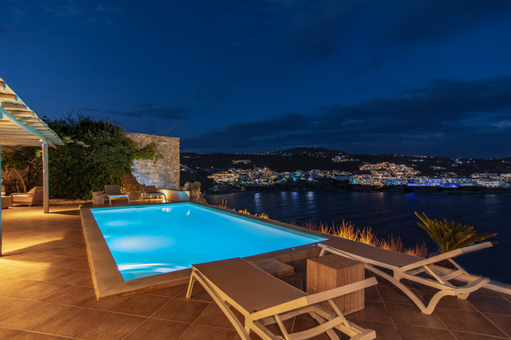 Lit pool overlooking coastal lights at night at Villa Patricia, for sale in Agios Lazaros, Mykonos